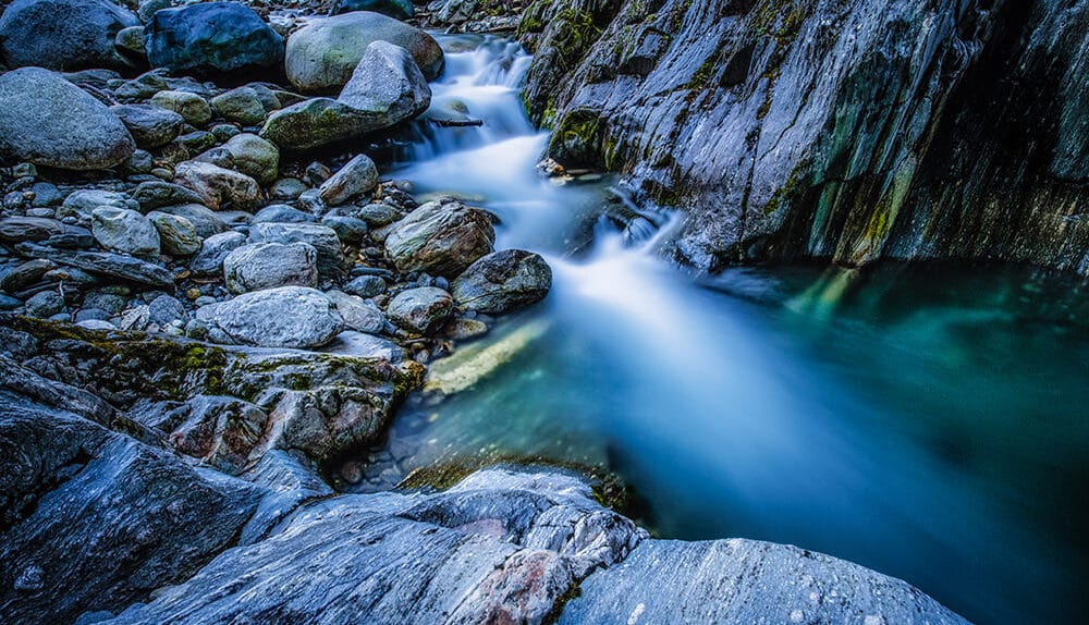 Langzeitbelichtung eines Bachs im Binntal, Wasser erscheint weich und verwischt zwischen Felsen und Steinen.