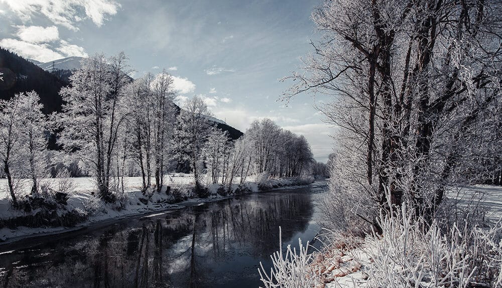 „Verschneite Winterlandschaft am Rotten im Oberwallis, mit vereisten Bäumen entlang des Flusses und Blick nach Westen unter leicht bewölktem Himmel.“